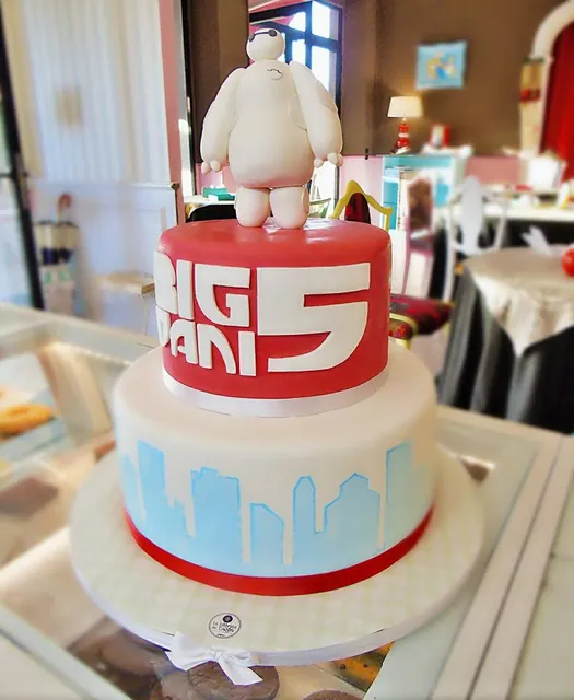 Torta a due piani con piano rosso, scritta bianca, skyline azzurro e statuina di Baymax in cima