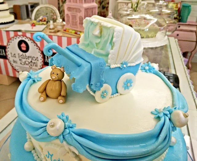 Primo piano di una torta azzurra con carrozzina di pasta di zucchero, orsetto, drappeggi e perle