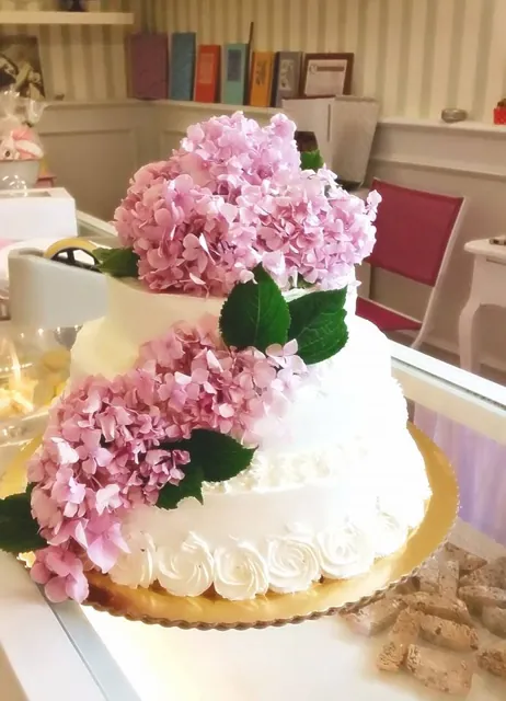 Torta bianca a più piani con rosette di panna alla base e una cascata di ortensie rosa e foglie verdi.