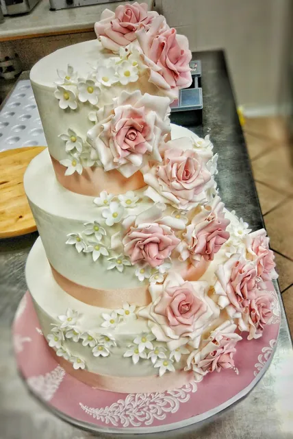 Torta a tre piani bianca con nastri pesca, cascata di rose rosa e fiorellini bianchi, base rosa con ricami a pizzo.