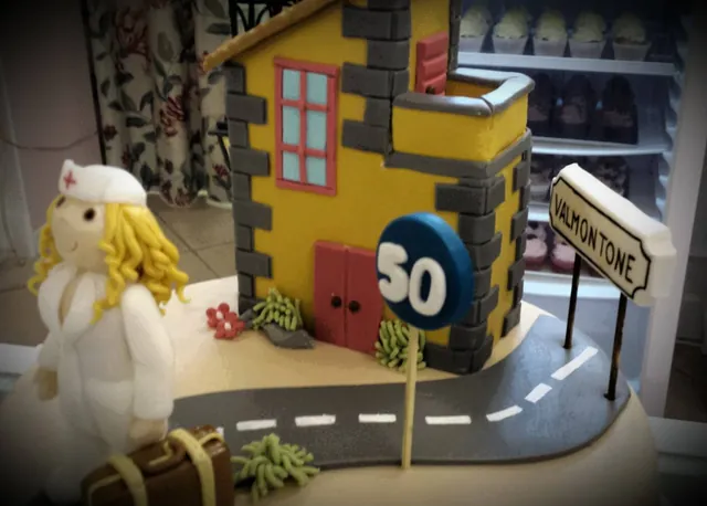 Torta scenografica con casetta gialla, strada, cartello con il numero 50 e figura di infermiera con valigia