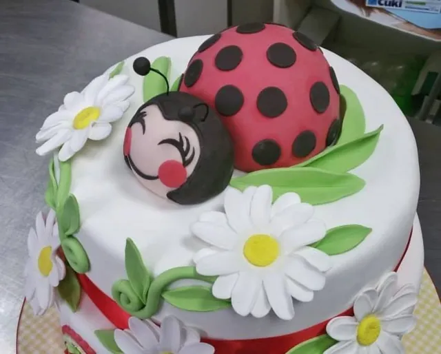 Primo piano di una coccinella di pasta di zucchero con guance rosa tra margherite e foglie verdi su una torta bianca