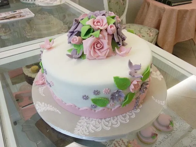 Torta rotonda bianca con mazzo di rose rosa e lilla, foglie verdi e fiorellini di zucchero