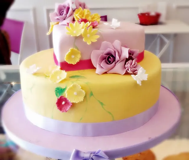 Torta a due piani con piano basso giallo, piano alto rosa, nastro fucsia, rose e margherite di zucchero e farfalle.