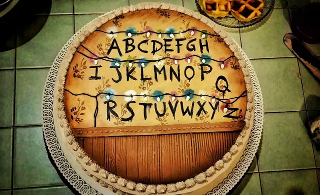 Torta tonda a tema serie TV con alfabeto dipinto su sfondo antico, lucine decorative e bordo in panna