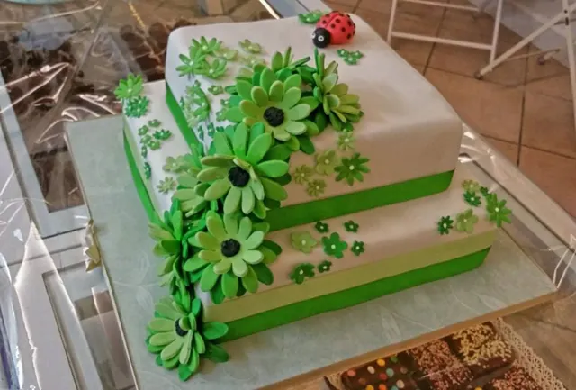 Torta quadrata a due piani grigio chiaro con una cascata di margherite verdi di zucchero, nastro verde e una coccinella in cima.