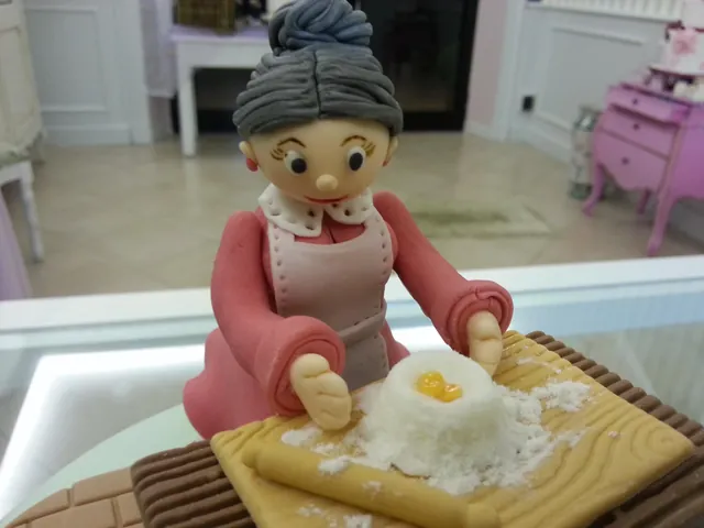 Statuina scultorea in pasta di zucchero di una nonna con grembiule che impasta farina e uovo su un tagliere, sopra una torta