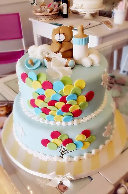 Torta azzurra a due piani decorata con palloncini colorati di zucchero e, in cima, un orsetto con biberon e ciuccio.