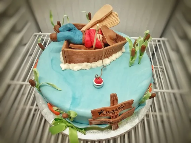 Torta tonda azzurra come un lago con barchetta di cioccolato, pescatore di pasta di zucchero, canne e cartelli di legno