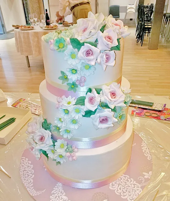 Torta nuziale a tre piani bianca con rose rosa, fiorellini azzurri e verde in cascata, con nastri rosa e decori a pizzo.