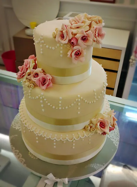 Torta nuziale a due piani color crema con festoni di perline, nastri e cascata di rose rosa in pasta di zucchero, su alzata trasparente.
