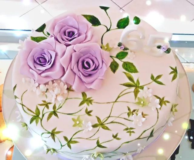 Torta rotonda in pasta di zucchero con tre rose viola, rami e foglioline dipinti a mano e un grande numero 65 sul bordo.