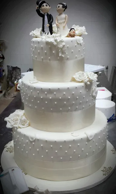 Torta nuziale a tre piani bianchi con perline, nastri e statuina degli sposi sulla sommità