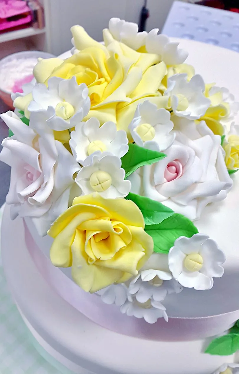 Primo piano di rose gialle e bianche e fiorellini modellati in pasta di zucchero sulla sommità di una torta