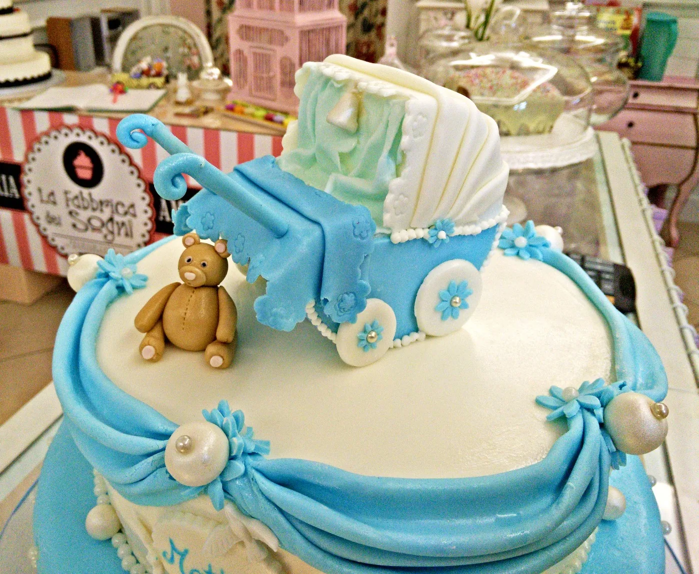 Primo piano di una torta azzurra con carrozzina di pasta di zucchero, orsetto, drappeggi e perle