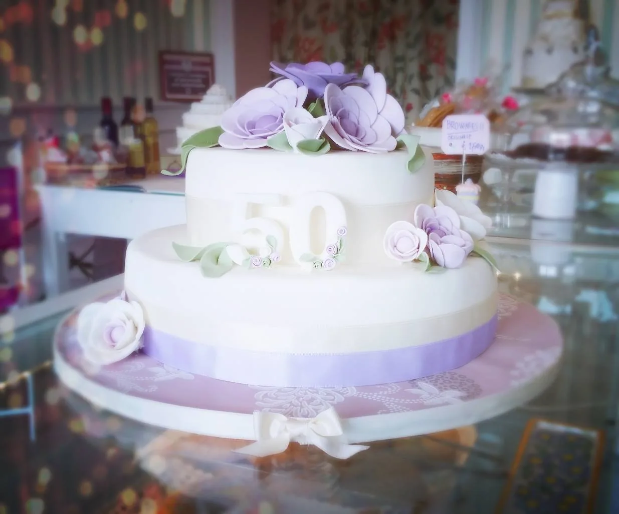 Torta bianca a due piani con grandi rose lilla di zucchero, un numero 50 in rilievo e nastro lilla su base rosa.