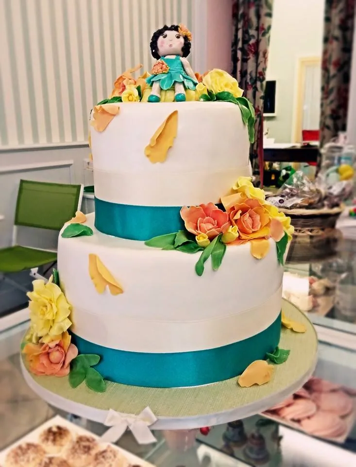 Torta a due piani bianca con nastro turchese, rose arancio e gialle e una bambolina in abito turchese in cima