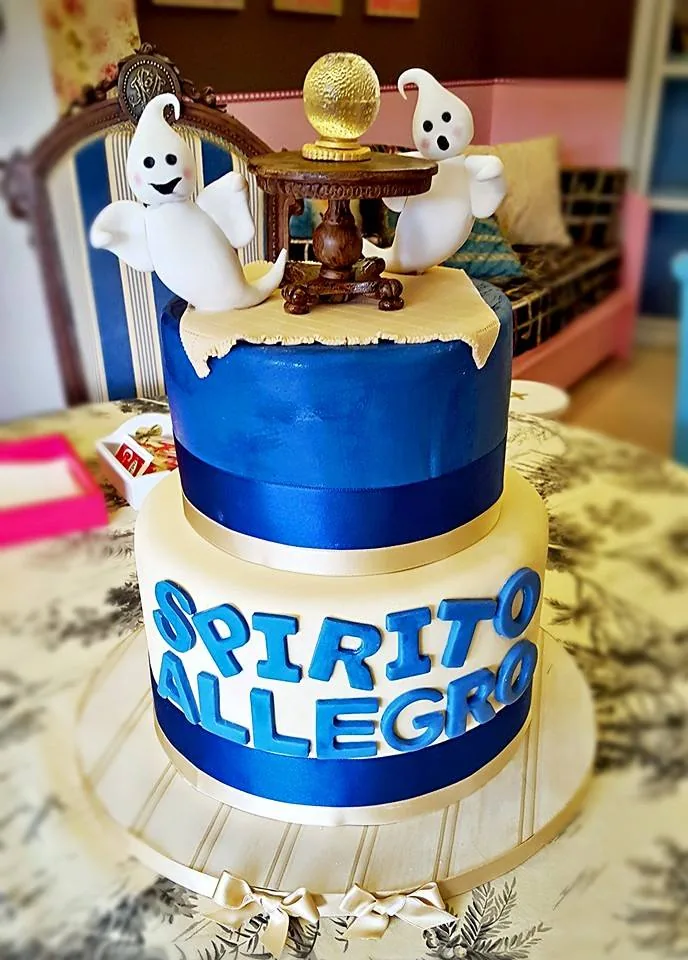 Torta a due piani blu e crema con lettere azzurre e due fantasmini bianchi attorno a un tavolino con sfera dorata