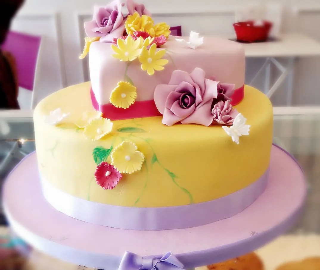 Torta a due piani con piano basso giallo, piano alto rosa, nastro fucsia, rose e margherite di zucchero e farfalle.