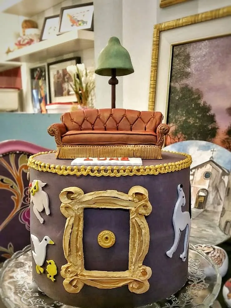 Torta rotonda viola scuro con divano di pasta di zucchero, lampada, cornice dorata attorno allo spioncino e animaletti bianchi