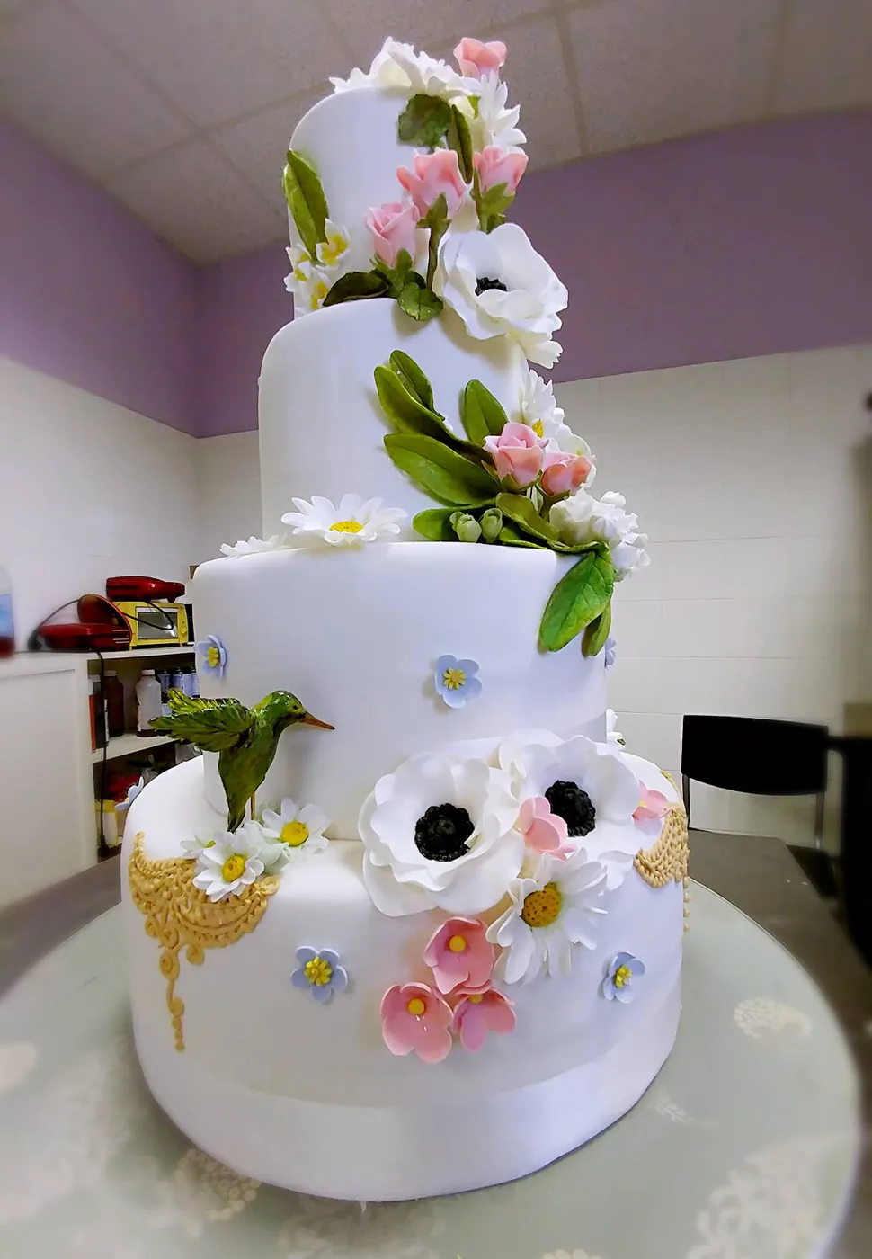 Torta nuziale bianca a quattro piani con anemoni, margherite, boccioli rosa, foglie verdi e un colibrì verde, ricami dorati alla base.