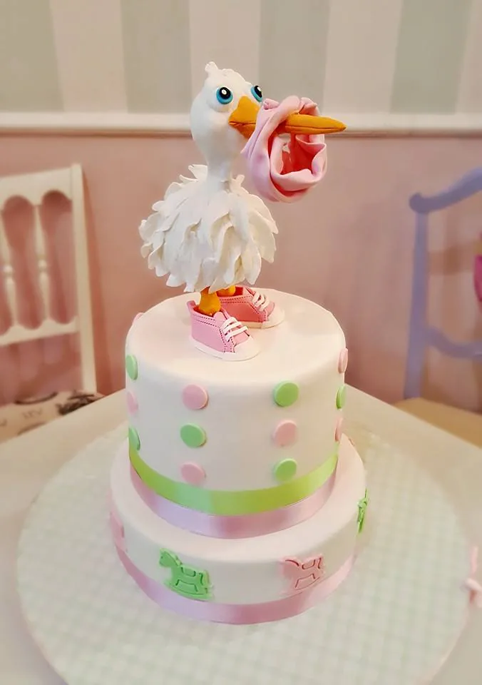 Torta a due piani bianca con pois rosa e verdi, cicogna di zucchero con fagotto rosa e scarpine rosa sulla sommità