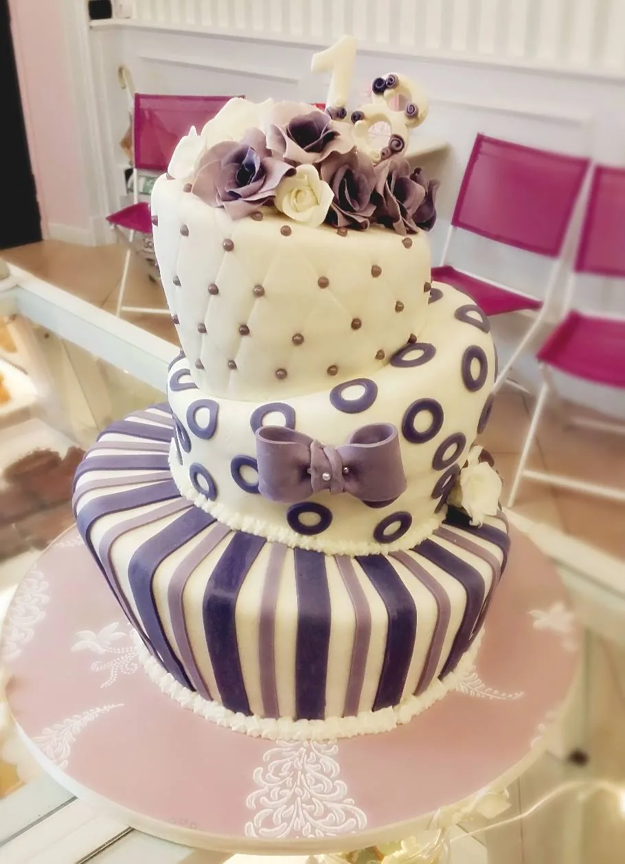 Torta a tre piani sfalsati, in stile wonky, con strisce viola, pois, fiocco grigio e rose di zucchero in cima, su base rosa.