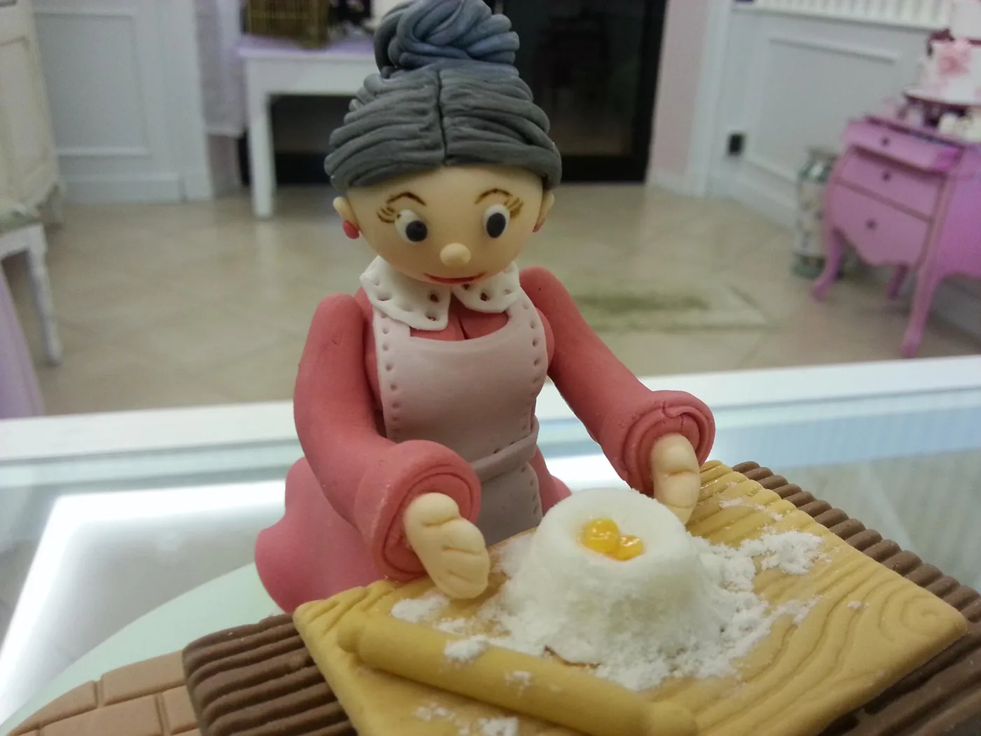 Statuina scultorea in pasta di zucchero di una nonna con grembiule che impasta farina e uovo su un tagliere, sopra una torta