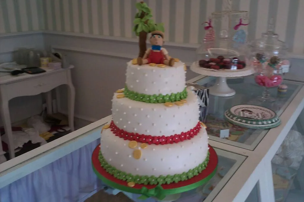 Torta a tre piani bianca con bordi rossi e verdi e statuina di Pinocchio con monete d'oro