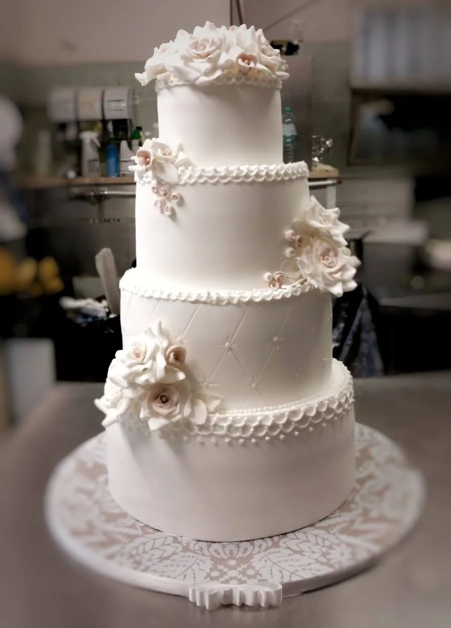 Torta nuziale bianca a quattro piani con rose di zucchero color cipria, perline di glassa e base a pizzo