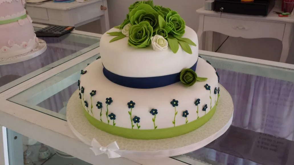 Torta a due piani bianca con rose verdi, nastro blu e fiorellini blu sul bordo