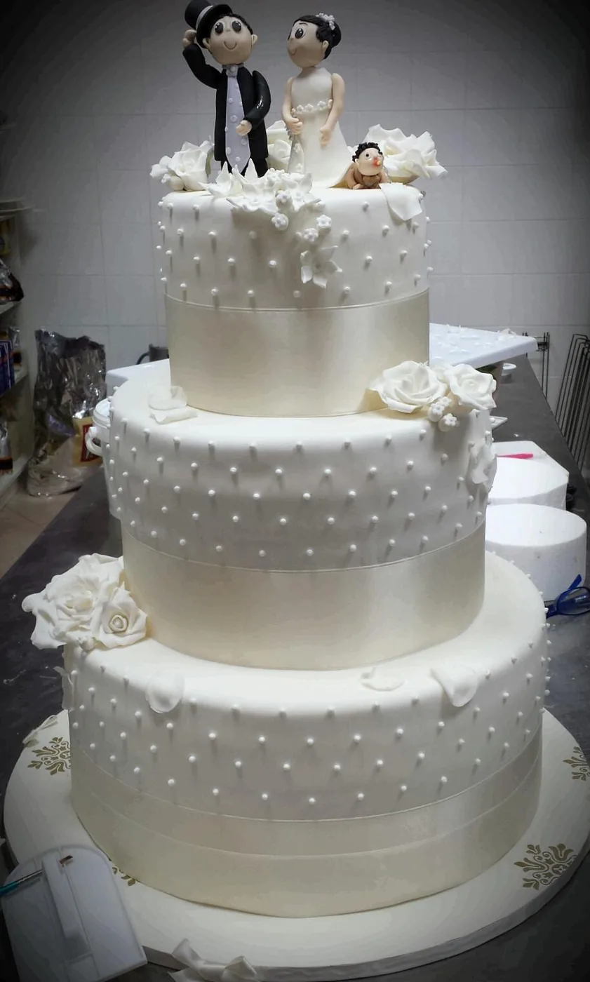 Torta nuziale a tre piani bianchi con perline, nastri e statuina degli sposi sulla sommità