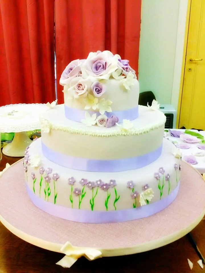 Torta a tre piani bianca e lilla con tulipani dipinti sul piano basso, nastri lilla e rose di zucchero in cima.