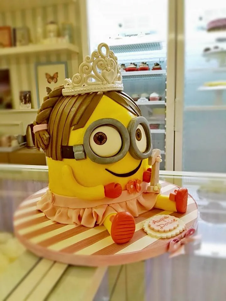 Torta scultura a forma di personaggio giallo con occhialoni, capelli castani, tiara bianca e gonnellina rosa su base a righe