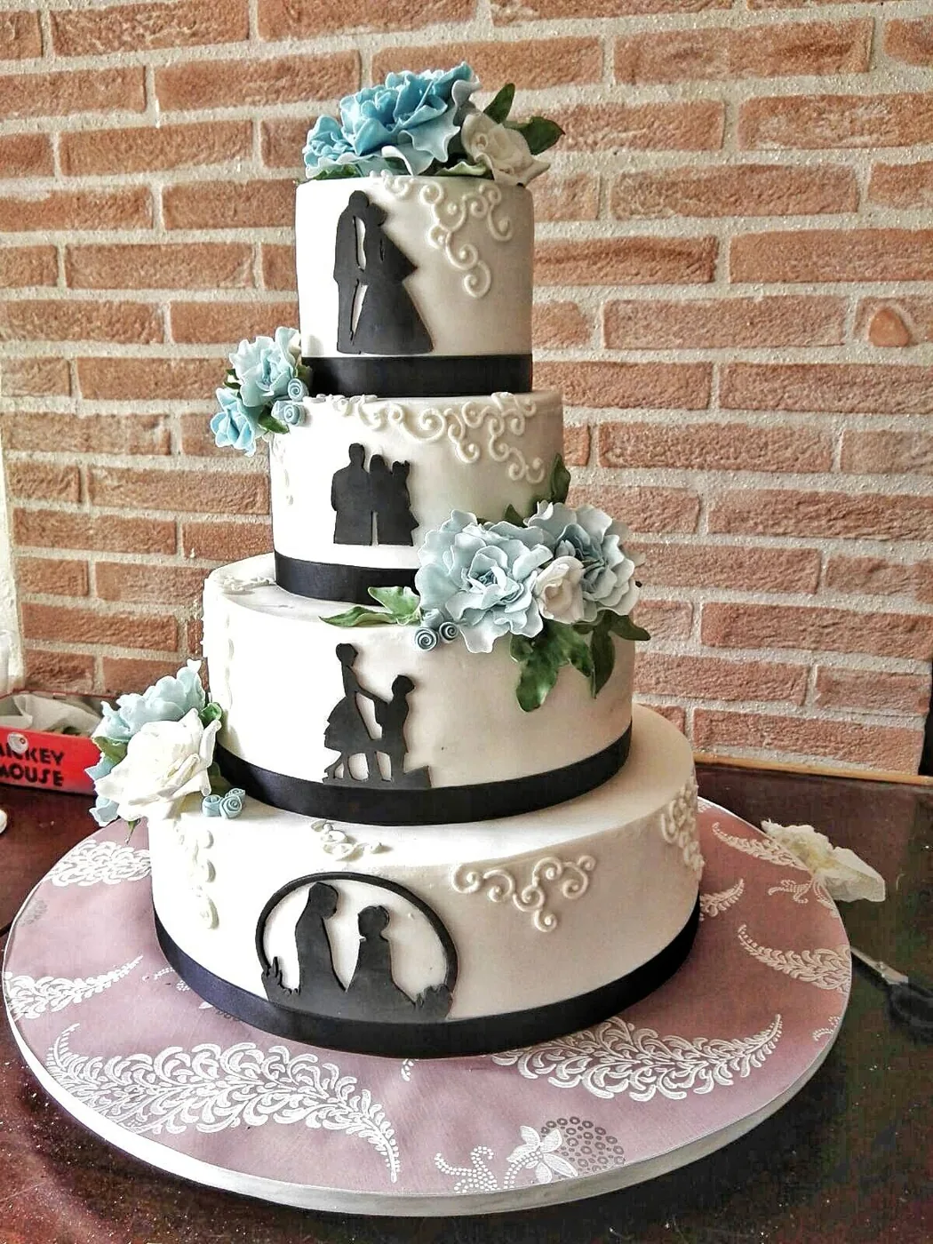 Torta nuziale a quattro piani bianca con silhouette nere degli sposi e peonie di zucchero azzurre, su fondo di mattoni